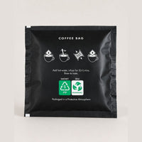 A single black coffee bag sachet with white printed text and icons, including coffee brewing steps and recycling symbols, photographed on a light neutral background.